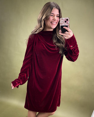 Woman wearing a burgundy velvet dress taking a mirror selfie on a green background