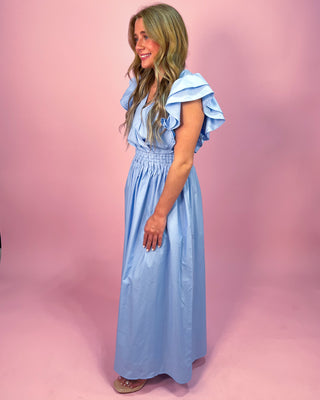 Woman wearing a light blue maxi dress with a wrap-style bodice, ruffled cap sleeves, and a smocked waistband, standing against a pink background.