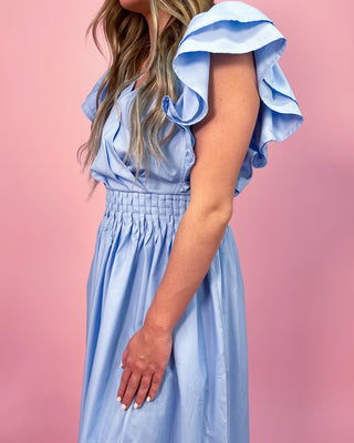 Woman wearing a light blue maxi dress with a wrap-style bodice, ruffled cap sleeves, and a smocked waistband, standing against a pink background.