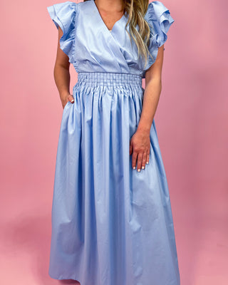 Woman wearing a light blue maxi dress with a wrap-style bodice, ruffled cap sleeves, and a smocked waistband, standing against a pink background.