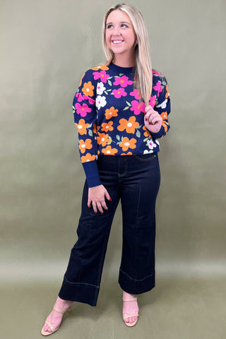 Woman wearing a colorful floral sweater and dark jeans against a plain background