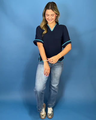 A woman modeling a navy blue short-sleeve top with turquoise trim, paired with light-wash jeans and metallic sneakers