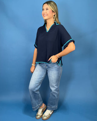 A woman modeling a navy blue short-sleeve top with turquoise trim, paired with light-wash jeans and metallic sneaker
