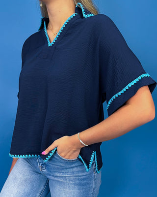 A woman wearing a navy blue short-sleeve blouse with bright turquoise blue scalloped trim, paired with light-wash blue jeans