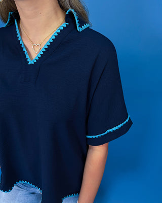 A close-up of a person wearing a navy blue short-sleeve top with bright turquoise blue scalloped trim along the V-neck and sleeve cuffs