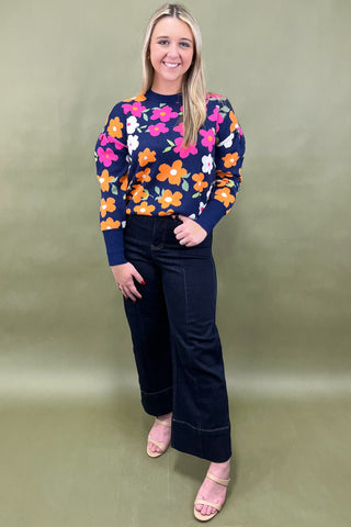 Woman wearing a colorful floral sweater and black pants against a green background