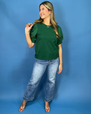 A woman modeling a dark green short-sleeve top with ruffled sleeves, paired with light-wash blue jeans and metallic sandals