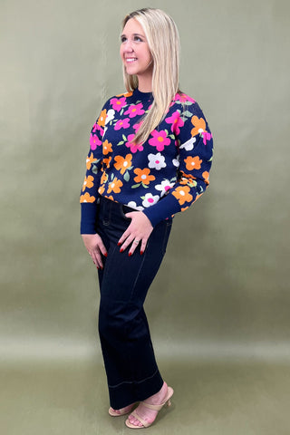Woman wearing a colorful floral sweater and dark pants against a plain background