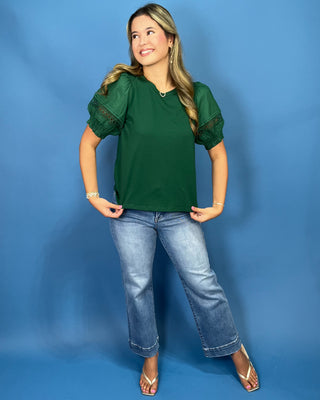 A woman modeling a dark green short-sleeve top with sheer lace puff sleeves, paired with light-wash blue jeans and white sandals
