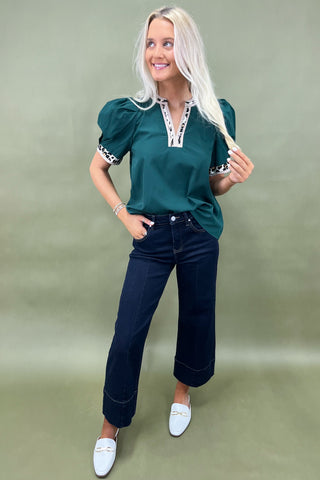 Woman wearing a green top with white embroidery and dark blue jeans on a plain background