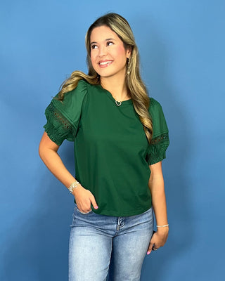 A woman modeling a dark green short-sleeve blouse with sheer puff sleeves and lace detailing at the cuffs, paired with light blue jeans