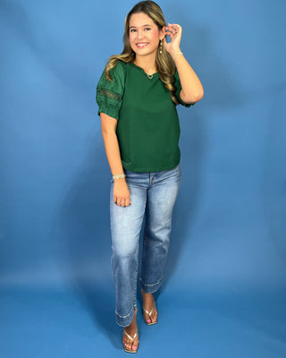A woman modeling a dark green short-sleeve blouse with sheer lace puff sleeves, paired with light-wash blue jeans and metallic sandals