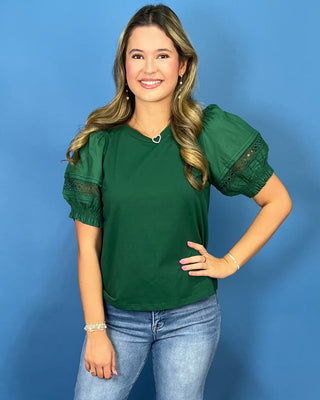 A woman modeling a dark green short-sleeve top with puff sleeves featuring lace trim, paired with light blue jeans