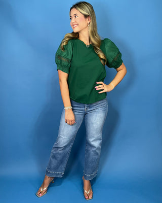A woman modeling a dark green short-sleeve blouse with sheer lace puff sleeves, paired with light-wash blue jeans and clear heeled sandals
