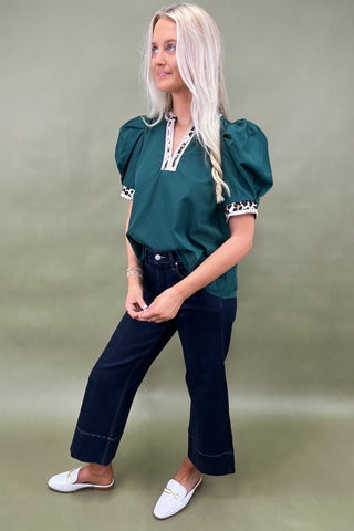 Woman wearing a green blouse with white trim and dark jeans against a plain background