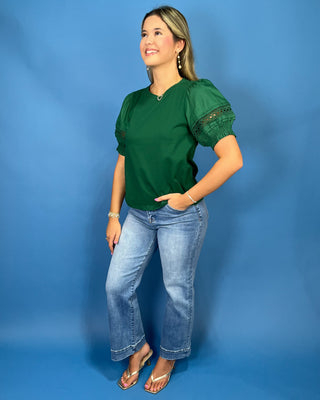 A woman modeling a dark green short-sleeve blouse with puff sleeves and lace detailing, paired with light-wash blue jeans and metallic sandals