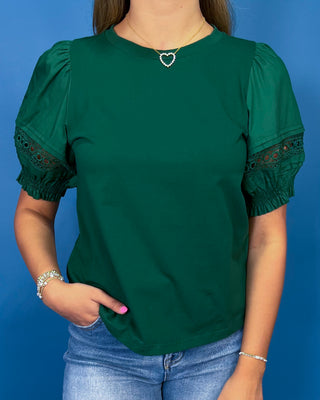 A woman wearing a forest green short-sleeve top with puff sleeves and lace trim at the cuffs, paired with blue jeans