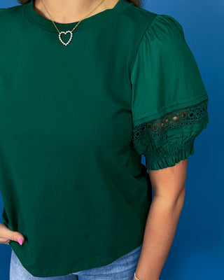 A woman wearing a dark green short-sleeve top with puff sleeves and black eyelet lace trim at the cuff