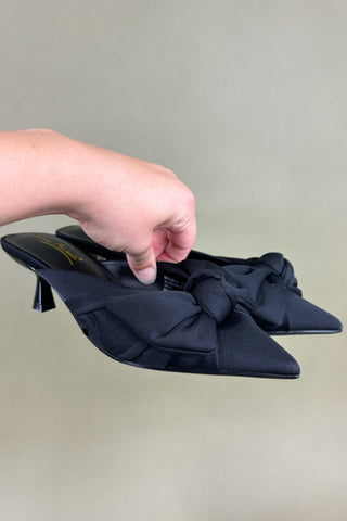 Black pointed-toe shoes with a bow held by a hand against a neutral background