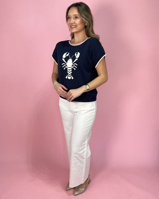 Woman wearing a navy t-shirt with a lobster graphic and white pants on a pink background