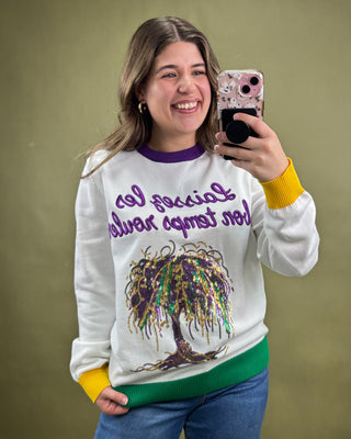 Woman wearing a colorful sweater with a tree design and text, taking a selfie against a plain background.
