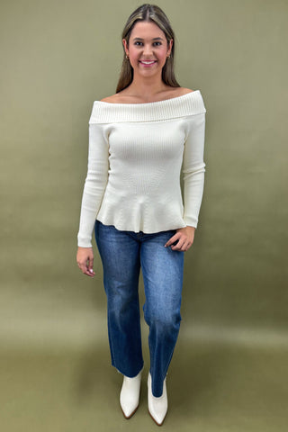 Woman wearing a white off-shoulder sweater and blue jeans against a green background