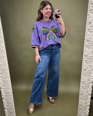 Woman taking a mirror selfie wearing a purple sweater with a colorful bow design and blue jeans.