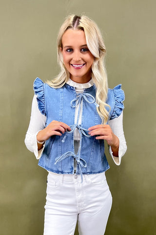Woman wearing a blue denim top with ruffled sleeves and white pants against a beige background