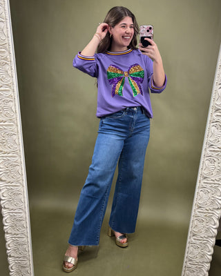 Person taking a mirror selfie wearing a purple sweatshirt with a colorful bow design and blue jeans.