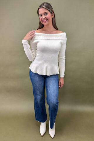 Woman wearing a white off-shoulder sweater and blue jeans against a plain background