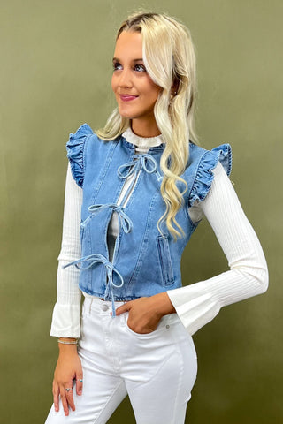 Woman wearing a blue denim vest with ruffles and white pants against a green background