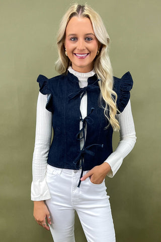 Woman wearing a navy blue vest with ruffled shoulders and a bow tie, standing against a green background.