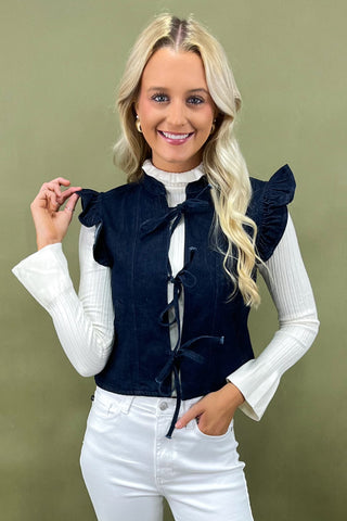 Woman wearing a navy blue vest over a white long-sleeve shirt with a green background