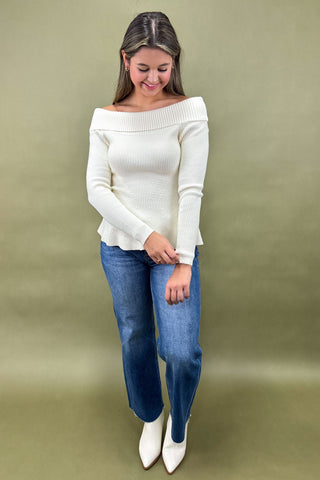 Woman wearing a white off-shoulder sweater and blue jeans against a beige background