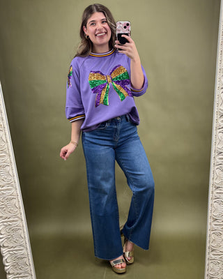 Woman taking a mirror selfie wearing a purple sweater with a colorful bow design and blue jeans.