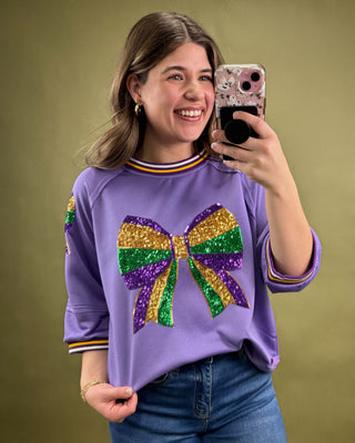 Person wearing a purple shirt with a colorful bow design, taking a selfie against a green background.