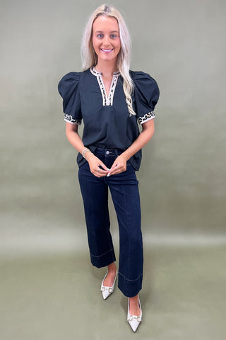 Woman wearing a black top with white accents and dark jeans on a plain background
