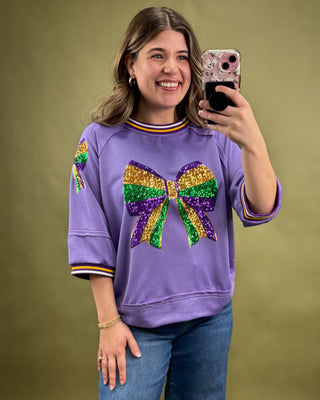Woman wearing a purple sweatshirt with a colorful bow design, taking a selfie against a green background.