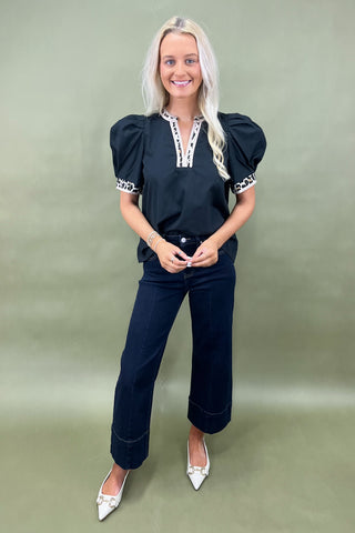 Woman wearing a black top with white accents and dark jeans on a plain background