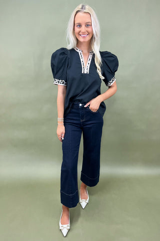 Woman wearing a black top with white accents and dark jeans on a plain background