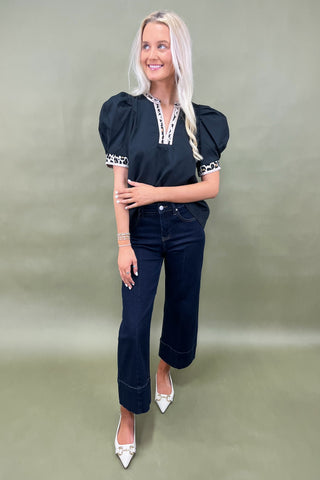 Woman wearing a black top with white accents and dark jeans on a green background