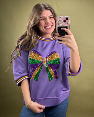 Woman taking a selfie wearing a purple sweatshirt with a colorful bow design on a green background