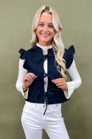 Woman wearing a navy blue top with ruffled shoulders and white pants against a green background