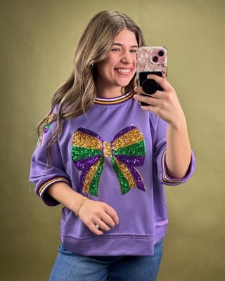 Person wearing a purple sweatshirt with a colorful bow design, taking a selfie against a green background.