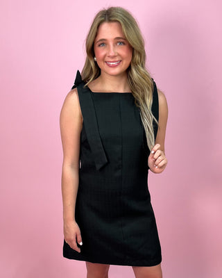 Woman wearing a black sleeveless mini dress with a square neckline and wide tie shoulder straps, paired with clear heeled sandals, standing against a pink background.