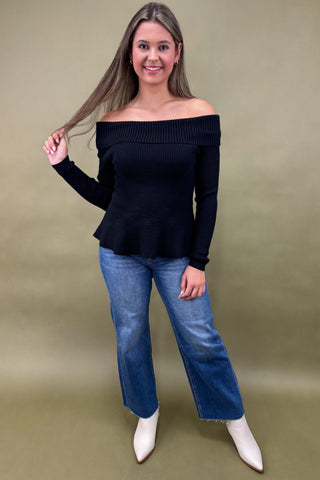 Woman wearing a black off-shoulder sweater and blue jeans against a beige background