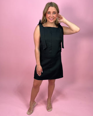 Woman wearing a black sleeveless mini dress with a square neckline and wide tie shoulder straps, paired with clear heeled sandals, standing against a pink background.