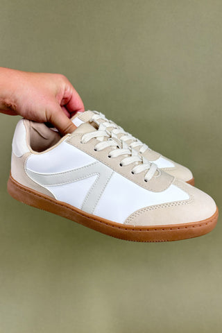 Beige and white sneakers with brown soles held by a hand against a green background