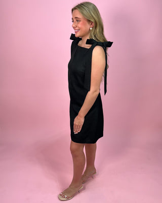 Woman wearing a black sleeveless mini dress with a square neckline and wide tie shoulder straps, paired with clear heeled sandals, standing against a pink background.