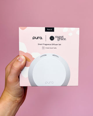 Hand holding a Pura smart fragrance diffuser packaging against a pink background